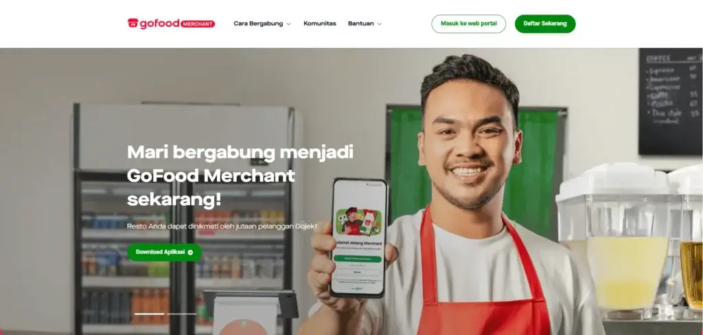 Tangkapan layar dari halaman beranda situs web resmi GoFood Merchant Indonesia. Di bagian atas halaman, terdapat logo merah "gofood MERCHANT" di sisi kiri, menu navigasi "Cara Bergabung", "Komunitas", "Bantuan", serta dua tombol aksi di sisi kanan yaitu "Masuk ke web portal" dan tombol hijau bertuliskan "Daftar Sekarang". Latar belakang utama gambar menampilkan suasana bagian dalam sebuah warung makan atau restoran, dengan kuliner pendingin minuman di sisi kiri dan dispenser jus di sisi kanan. Di bagian depan tengah, seorang pria pemilik warung makan tersenyum ramah mengenakan celemek merah khas GoFood, sambil memegang dan menunjukkan sebuah smartphone di tangan kanannya. Layar smartphone tersebut menampilkan antarmuka halaman awal aplikasi GoBiz atau GoFood Merchant dengan tulisan "Selamat datang Merchant" dan tombol "Daftar GoBiz sekarang". Di sebelah kiri pria tersebut, terdapat teks besar berwarna putih bertuliskan "Mari bergabung menjadi GoFood Merchant sekarang!" yang diikuti sub-teks "Resto Anda dapat dinikmati oleh jutaan pelanggan Gojek!" serta tombol hijau bertuliskan "Download Aplikasi". Visual ini mengomunikasikan dengan jelas bahwa cara daftar GoFood dapat diakses dan dilakukan sepenuhnya melalui aplikasi mobile.